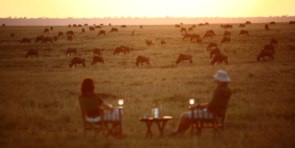 Two guests sit with drinks at sunset, watching a herd of wildebeest graze peacefully, bringing full circle the spirit behind frequently asked questions about safaris.
